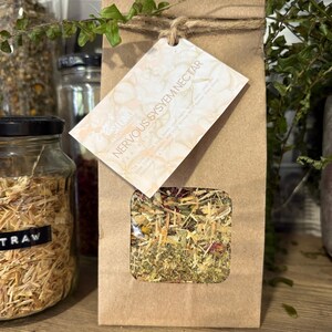 May include: A brown paper bag with a clear window showing a blend of dried herbs and flowers. A tag reads "NERVOUS SYSTEM NECTAR TEA BLEND". The bag is tied with twine and is next to glass jars.