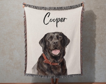 May include: A cream-colored blanket featuring a watercolor portrait of a black Labrador Retriever with the name "Cooper" in black script. The blanket has a fringed edge and the text "Watercolor Pet Portrait Blanket" at the top.