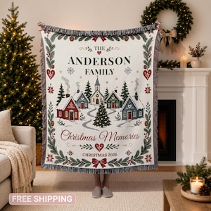 May include: A woven throw blanket featuring a cream background with a festive winter village scene, pine trees, and red hearts. The text reads THE ANDERSON FAMILY, Christmas Memories, and CHRISTMAS 2026. It has a fringed edge and decorative border.