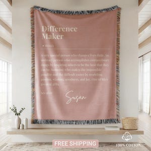 May include: A dusty rose woven throw blanket featuring the text Difference Maker, noun, followed by a definition describing a person who changes lives daily, and the name Susan in script. The blanket has fringed edges with multicoloured threads.