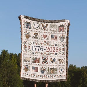 Peut inclure: Un plaid tissé avec un fond crème présentant une grille de motifs patriotiques et d'art populaire, incluant des maisons, un pygargue à tête blanche, la Liberty Bell, un drapeau américain, un mouton, un coq, une vache et le texte 1776-2026, avec des bords à franges.