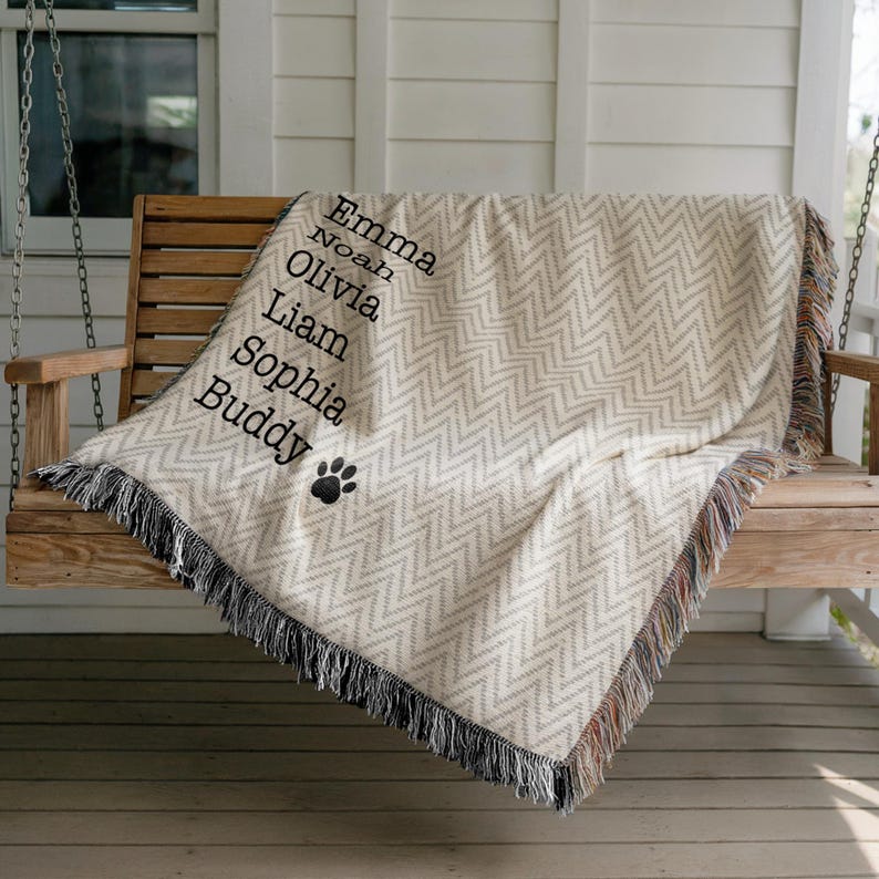 May include: A cream-colored throw blanket with a gray chevron pattern and black fringe. The blanket is personalized with names and a paw print. The names are Emma, Noah, Olivia, Liam, Sophia, and Buddy.