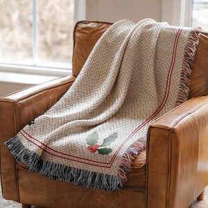 May include: A beige herringbone-patterned throw blanket with red trim and a holly design, draped over a brown leather armchair. The blanket has a fringed edge with black, red, and beige threads. The holly design features green leaves and red berries.