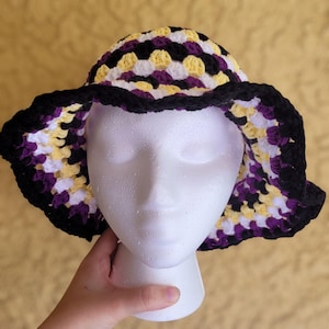May include: A handmade crochet bucket hat in a non-binary pride flag color scheme. The hat features a black brim and a crown with a pattern of yellow, white, and purple squares. The hat is displayed on a white mannequin head.