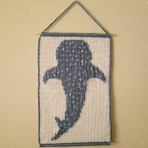 May include: A handmade, wall-hanging textile featuring a crocheted whale shark design. The whale shark is depicted in shades of blue and white, with a textured appearance. The piece is rectangular, with a wooden dowel at the bottom and top for hanging.
