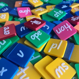 May include: A close-up of a pile of colourful square tiles, each bearing white letters or letter combinations. The tiles are in shades of blue, green, yellow, pink, and red. Some tiles show words such as "tion", "augh", and "ate".