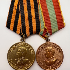 May include: Two circular bronze-toned medals featuring a profile portrait of a man in military uniform. Each medal has the Cyrillic text "НАШЕ ДЕЛО ПРАВОЕ МЫ ПОБЕДИЛИ" inscribed along the edge. They are attached to ribbons with orange and black stripes.