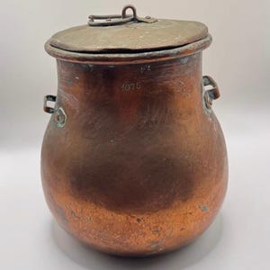 Könnte beinhalten: Ein antiker Kupfertopf mit Deckel. Der Topf hat einen abgerundeten Körper, zwei seitliche Griffe und einen Klappdeckel mit Schlaufengriff. Die Nummer "1075" ist auf der Seite eingraviert. Der Topf hat ein verwittertes Aussehen.