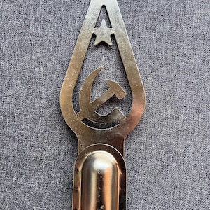 May include: A metallic spearhead-shaped finial featuring a cutout design of a five-pointed star positioned above a hammer and sickle symbol. The base of the object consists of a cylindrical socket designed for attachment to a pole or staff.
