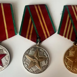 May include: Three medals with red and yellow striped ribbons. The medals have star designs, one with a red star and hammer and sickle, one with a gold star, and one with a silver star.