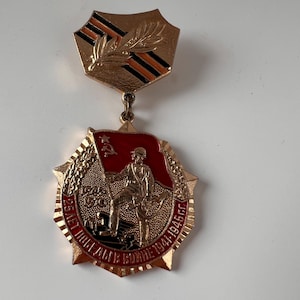 Può includere: Una medaglia commemorativa color oro raffigurante un soldato che regge una bandiera con il simbolo di falce e martello. La medaglia presenta dettagli in smalto rosso, un bordo testurizzato a forma di stella e una barra di sospensione.