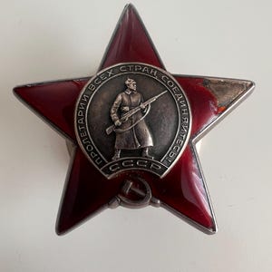 Puede incluir: Una medalla en forma de estrella de cinco puntas con un acabado de esmalte rojo oscuro y un borde plateado. En el centro, un medallón circular muestra a un soldado con un rifle, rodeado de texto cirílico y el acrónimo CCCP abajo.
