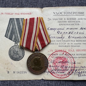 May include: A Soviet Medal for the Victory over Japan with a red, white, and yellow striped ribbon, placed on a document titled UDOSTOVERENIE. The medal features a profile portrait of Joseph Stalin and Cyrillic text, with serial number 162234.