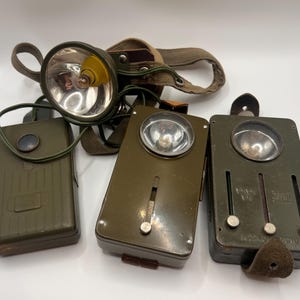 Könnte beinhalten: Eine Sammlung von olivgrünen Vintage-Stirnlampen und Akkus. Eine Stirnlampe hat ein kreisförmiges Licht mit einer gelben Glühbirne und einem braunen Riemen. Die anderen drei sind rechteckig mit kreisförmigen Lichtern und Knöpfen.