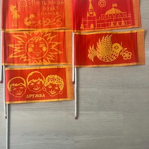 Puede incluir: Conjunto de cinco banderas de tela roja con ilustraciones amarillas. Las banderas representan un sol, un pájaro, un edificio, un niño jugando y tres niños con la palabra "DRUZHBA" escrita debajo.