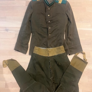 Op de afbeelding: Een olijfgroen militair uniform bestaande uit een jas en een broek. De jas heeft een hoge kraag met gouden en groene accenten en gouden schouderstukken. De broek is donkergroen met een lichtere groene tailleband en beenuiteinden.