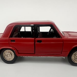 Puede incluir: Un coche modelo rojo fundido a presión con cuatro puertas y un interior detallado. El coche tiene ruedas doradas y parachoques negros. El modelo es un diseño de sedán clásico, probablemente un vehículo vintage o retro.