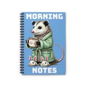 Peut inclure: Cahier à spirales avec une couverture bleue. Il présente un opossum de dessin animé en peignoir vert, tenant une tasse et portant des pantoufles en forme de lapin. Les mots "MORNING NOTES" sont imprimés en blanc.