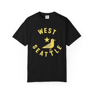 May include: Black t-shirt with a yellow graphic featuring a seagull, star, and the words "WEST SEATTLE". The design is centered on the front of the shirt. The shirt has short sleeves and a crew neck.