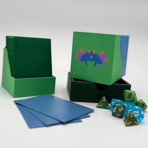May include: A green card holder with a stack of green cards, a green and blue box with a colorful design, a small black box, blue cards, and several dice. The dice are green and blue.