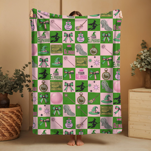 Peut inclure: Une couverture à carreaux roses et verts avec des motifs de sorcières. Le motif comprend des chapeaux de sorcières, des nœuds, des silhouettes de sorcières sur des balais et les textes "Team Elphaba" et "Team Glinda".