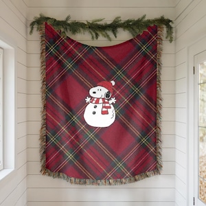 Puede incluir: Una manta a cuadros rojos con un diseño de muñeco de nieve Snoopy. La manta tiene un borde con flecos y presenta un muñeco de nieve Snoopy con un gorro y bufanda rojos. El patrón de cuadros incluye líneas verdes y amarillas.