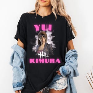 May include: Black t-shirt featuring a graphic of a woman with the text "Yui Kimura" in pink, along with a symbol. The shirt is a casual style, with rolled-up sleeves.