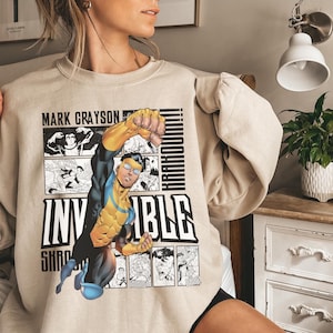 Könnte beinhalten: Beiges Sweatshirt mit einem Comic-Grafik eines Superhelden in Aktion. Das Design enthält die Wörter "MARK GRAYSON" und "INVINCIBLE" in schwarzer, fetter Schrift, Comic-Panels und das Wort "KRAKOOM!!".