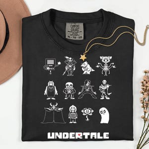 May include: Black t-shirt with white cartoon character illustrations from the video game Undertale. The shirt has a crew neck and a Comfort Colors tag. A gold necklace with a star pendant is draped across the shirt.