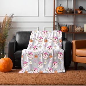 May include: A white throw blanket with a repeating pattern of cartoon ghosts wearing pink cowboy hats, bandanas, and holding lassos. The design also includes orange pumpkins, white daisies, and pink bows. The blanket is draped over a black leather couch.