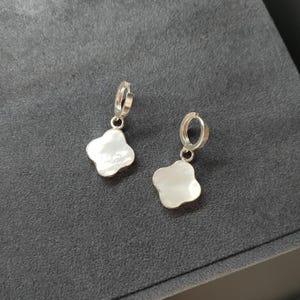 Peut inclure: Une paire de boucles d'oreilles créoles argentées avec un pendentif en forme de trèfle à quatre feuilles. Le pendentif a une surface blanche et irisée. Les boucles d'oreilles sont présentées sur une surface gris foncé.