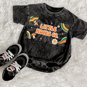 Op de afbeelding: Een zwarte baby romper met een vintage-stijl grafische afbeelding die "Little Mocks Co." zegt in regenboogkleuren. De romper is gemaakt van een zacht, comfortabel materiaal en heeft een drukknoopsluiting.