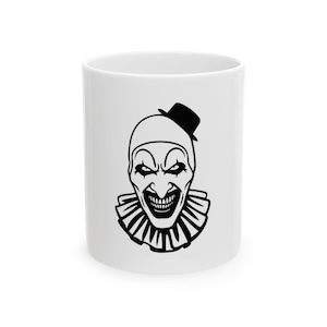 Peut inclure: Un mug en céramique blanche orné d'une illustration graphique noire représentant un visage de clown. Le clown porte un chapeau pointu, affiche un large sourire avec des dents visibles et porte une collerette plissée sur le mug cylindrique.