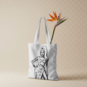 May include: A white tote bag featuring a black and white graphic illustration of a person with long hair wearing a sleeveless dress. A single orange and blue bird of paradise flower is placed inside the bag, extending upwards from the opening. This image is part of the RuPaul Drag Race Clipart Bundle, Queen of Drag SVG PNG JPG digital download.