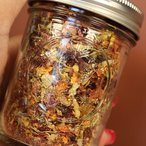 Könnte beinhalten: Ein klares Glas mit getrockneten Kräutern und Blumen, gehalten von einer Hand. Das Glas hat einen silbernen Deckel und geprägte Blumendesigns. Der Inhalt ist eine Mischung aus Orange, Gelb und Braun.