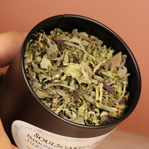 May include: A black tin filled with a herbal blend. The tin has a white label with the text "SOULSOAKED IMMUNITY SPICE HERBAL BLEND". The herbs are a mix of green and purple leaves.