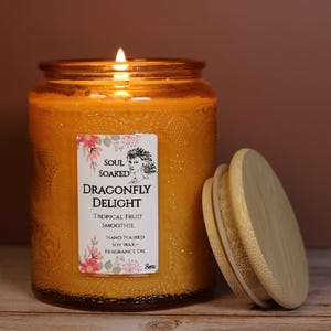 May include: An orange candle in a glass jar with a wooden lid. The label reads "Dragonfly Delight" and "Tropical Fruit Smoothie." The candle is lit, with a flame. The candle sits on a wooden surface.