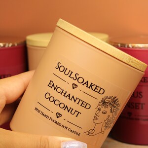 Può includere: Una candela di soia color pesca con coperchio in legno, etichettata "SOULSOAKED ENCHANTED COCONUT". L'etichetta presenta un motivo a diamante e un disegno a tratto del volto di una donna. La candela è da 295 ml.