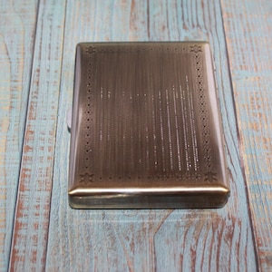 May include: A rectangular, silver-toned cigarette case with a textured surface and decorative border. The case is closed and sits on a weathered, blue and brown wooden surface.
