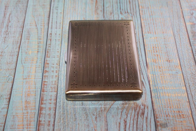 May include: A rectangular, silver-toned cigarette case with a textured surface and decorative border. The case is closed and sits on a weathered, blue and brown wooden surface.