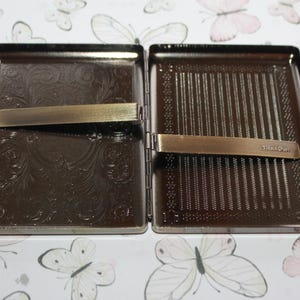 Puede incluir: Una pitillera rectangular de metal con apertura abatible y acabado en bronce oscuro. El interior presenta dos patrones en relieve distintos: un dise&ntilde;o de volutas florales a la izquierda y rayas verticales a la derecha. Un clip de metal est&aacute; sujeto a cada lado, con la palabra TitanOwl grabada en el clip derecho.