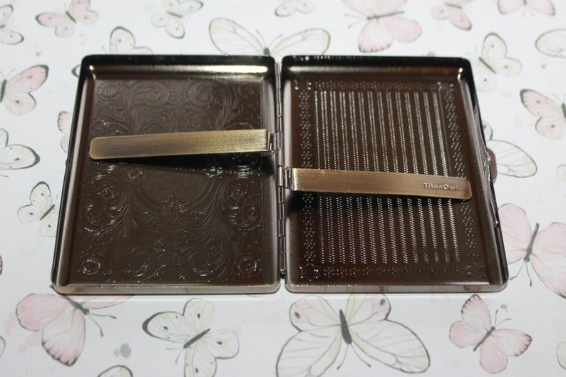 Puede incluir: Una pitillera rectangular de metal con apertura abatible y acabado en bronce oscuro. El interior presenta dos patrones en relieve distintos: un dise&ntilde;o de volutas florales a la izquierda y rayas verticales a la derecha. Un clip de metal est&aacute; sujeto a cada lado, con la palabra TitanOwl grabada en el clip derecho.