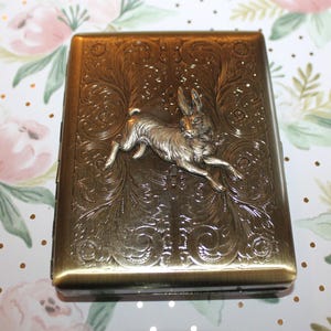 May include: A rectangular, antique brass-colored case with an embossed floral design and a silver-colored rabbit in mid-leap. The case has a hinged lid and a clasp closure. The background is a floral pattern.
