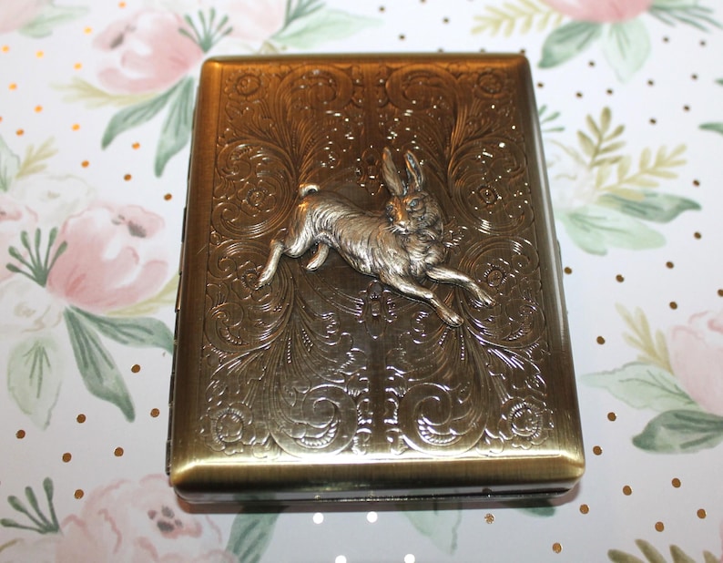 May include: A rectangular, antique brass-colored case with an embossed floral design and a silver-colored rabbit in mid-leap. The case has a hinged lid and a clasp closure. The background is a floral pattern.