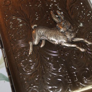 May include: A rectangular, antique-style case with a silver-colored rabbit figurine. The case has an ornate, embossed floral design and a dark bronze finish. The rabbit is in mid-leap, adding a dynamic element to the design.
