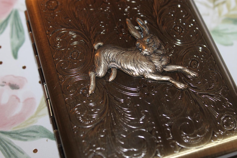 May include: A rectangular, antique-style case with a silver-colored rabbit figurine. The case has an ornate, embossed floral design and a dark bronze finish. The rabbit is in mid-leap, adding a dynamic element to the design.