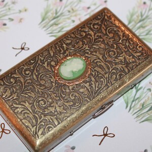 May include: A gold metal cigarette case with an ornate floral design. The case has a hinged lid and a small green cameo with a white silhouette in the center.