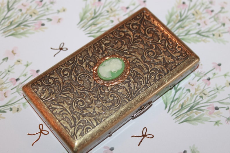 May include: A gold metal cigarette case with an ornate floral design. The case has a hinged lid and a small green cameo with a white silhouette in the center.