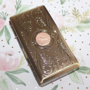 May include: A rectangular, antique-style metal case with an ornate floral design and a circular emblem featuring a leaping animal. The case is bronze-colored and has a hinged closure. The background is a floral pattern.