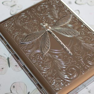 May include: A silver-colored rectangular case with a detailed dragonfly design on the top. The dragonfly is in the center, with intricate floral patterns surrounding it. The case has a shiny, metallic finish.